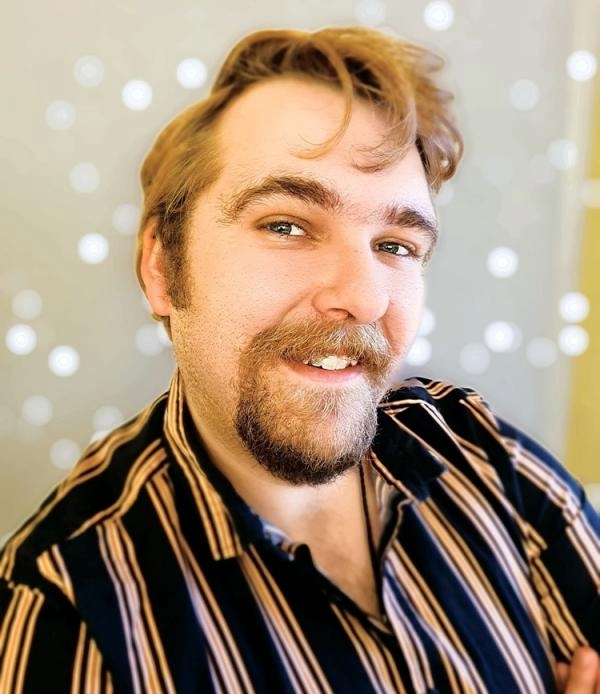 Man with beard in striped shirt with string lights in background.