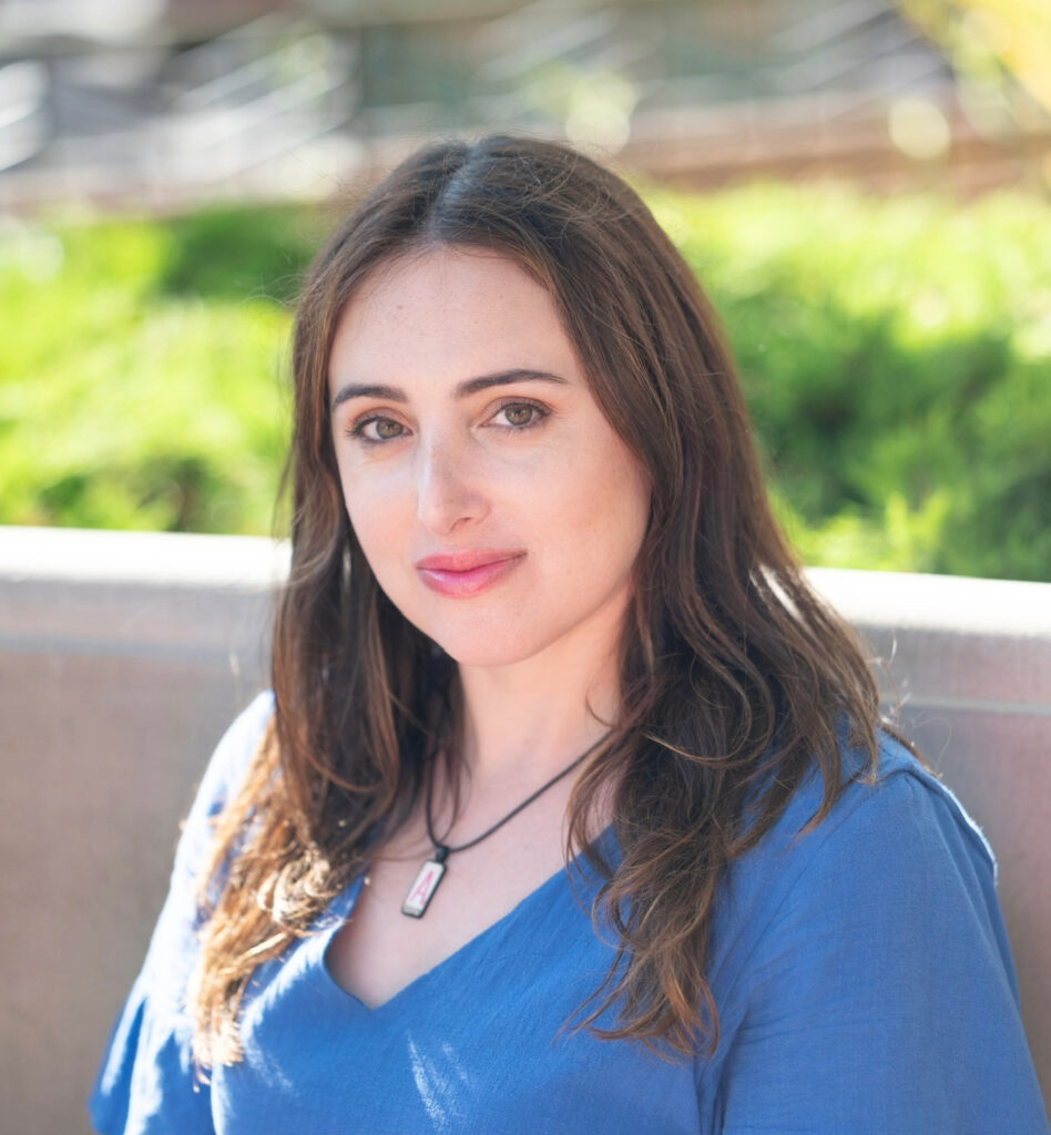 Woman wearing blue shirt and necklace, with blurred nature background.